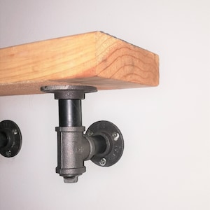 May include: A wooden shelf with black metal pipe brackets. The shelf is supported by two brackets, each with a round flange and a T-shaped pipe.