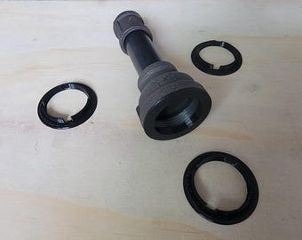 Może przedstawiać: Czarny metalowy element łączący rury z trzema czarnymi plastikowymi pierścieniami. Element łączący rury ma gwintowany koniec i gładkie, cylindryczne ciało. Pierścienie mają średnicę około 5 cm i mają niewielką szczelinę.