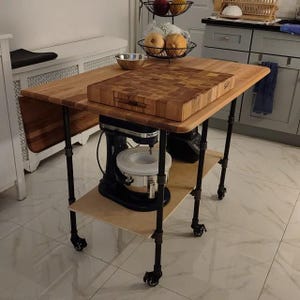 May include: A kitchen island cart with a light wood top, a thick butcher block cutting board, and a black metal pipe frame with four caster wheels. A black stand mixer with a stainless steel bowl sits on the lower shelf of the cart.