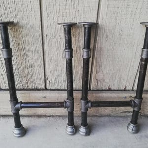 May include: Two sets of dark gray metal pipe furniture legs, each featuring two vertical pipes connected by a horizontal crossbar. The legs have circular mounting plates at the top and rounded end caps at the bottom, constructed from industrial-style threaded pipe fittings.