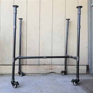 May include: Black metal industrial pipe frame with four wheels. The frame is made of black metal pipes and has four wheels on the bottom. The frame is standing on a concrete surface.