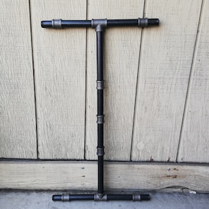 May include: A black metal T-shaped pipe fitting with silver-colored connectors. The pipe fitting is mounted on a wall.