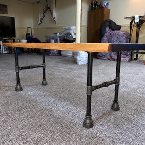 May include: A rectangular table featuring a natural wood slab top with a live edge and a dark brown finish. The base is constructed from black industrial metal pipes and fittings, including four vertical legs connected by horizontal support bars.