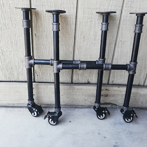 May include: Four black metal table legs with adjustable feet and casters. The legs are made of industrial pipe and have a black finish. The casters are black and have a silver accent.