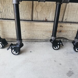 May include: Four black metal legs with black wheels on a concrete surface. The legs are attached to a metal frame.
