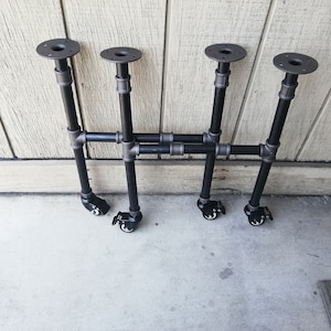 May include: Four black metal table legs with wheels. Each leg has a round metal plate at the top and is made of pipe sections. The legs are arranged in two pairs, with the pairs connected by a horizontal pipe section.