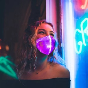 Puede incluir: Una mujer con una máscara facial LED de luz púrpura. La máscara tiene un patrón geométrico y está iluminada con luces moradas brillantes. La mujer está de pie frente a un letrero de neón.