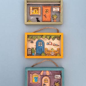 May include: Three framed miniature scenes of buildings, each with a different color frame and a rope for hanging. The top frame is green and depicts a workshop. The middle frame is yellow and shows a cafe. The bottom frame is teal and shows a tailor shop.