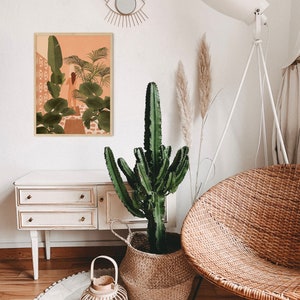 Puede incluir: Una silla de mimbre blanca con una gran planta de cactus verde en una cesta tejida. El cactus est&aacute; delante de una c&oacute;moda blanca con dos cajones. Una impresi&oacute;n enmarcada de una mujer en un entorno tropical cuelga de la pared sobre la c&oacute;moda. Una l&aacute;mpara blanca con una pantalla blanca est&aacute; en el fondo.