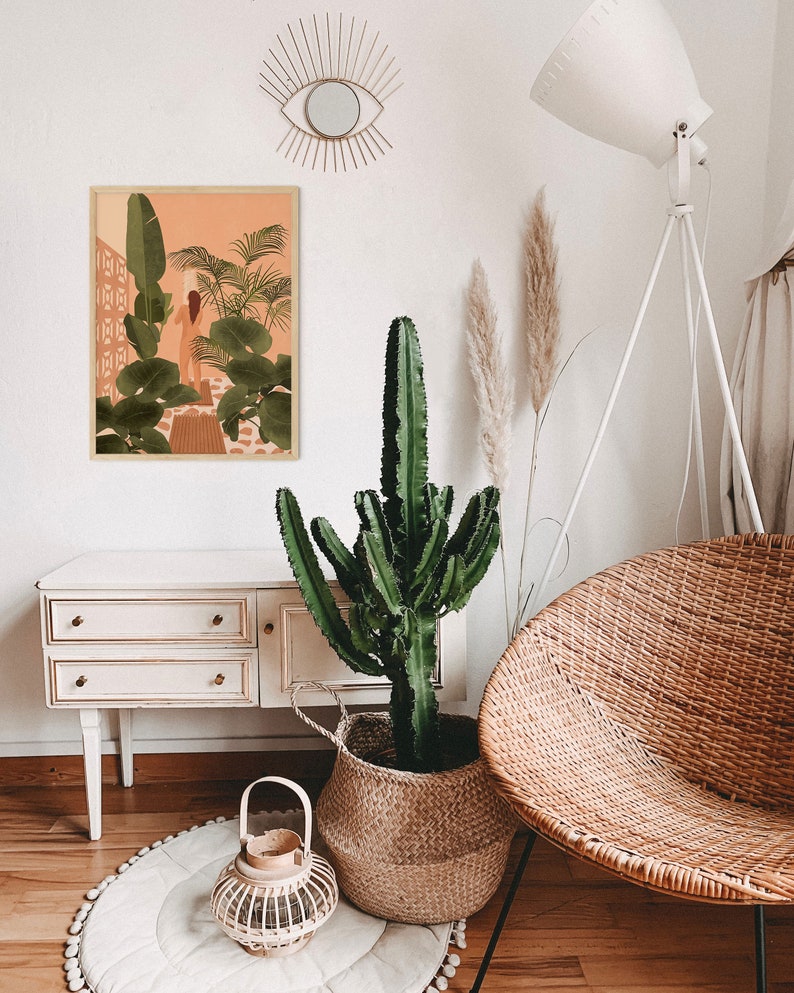 Puede incluir: Una silla de mimbre blanca con una gran planta de cactus verde en una cesta tejida. El cactus est&aacute; delante de una c&oacute;moda blanca con dos cajones. Una impresi&oacute;n enmarcada de una mujer en un entorno tropical cuelga de la pared sobre la c&oacute;moda. Una l&aacute;mpara blanca con una pantalla blanca est&aacute; en el fondo.