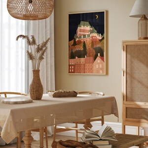 May include: A beige tablecloth covers a wooden dining table with two chairs. A brown throw blanket is draped over a wooden bench with a stack of books. A framed illustration of a city skyline with a crescent moon hangs on the wall above the table. A wicker lampshade hangs from the ceiling above the table. A wicker vase with dried flowers sits on the table.