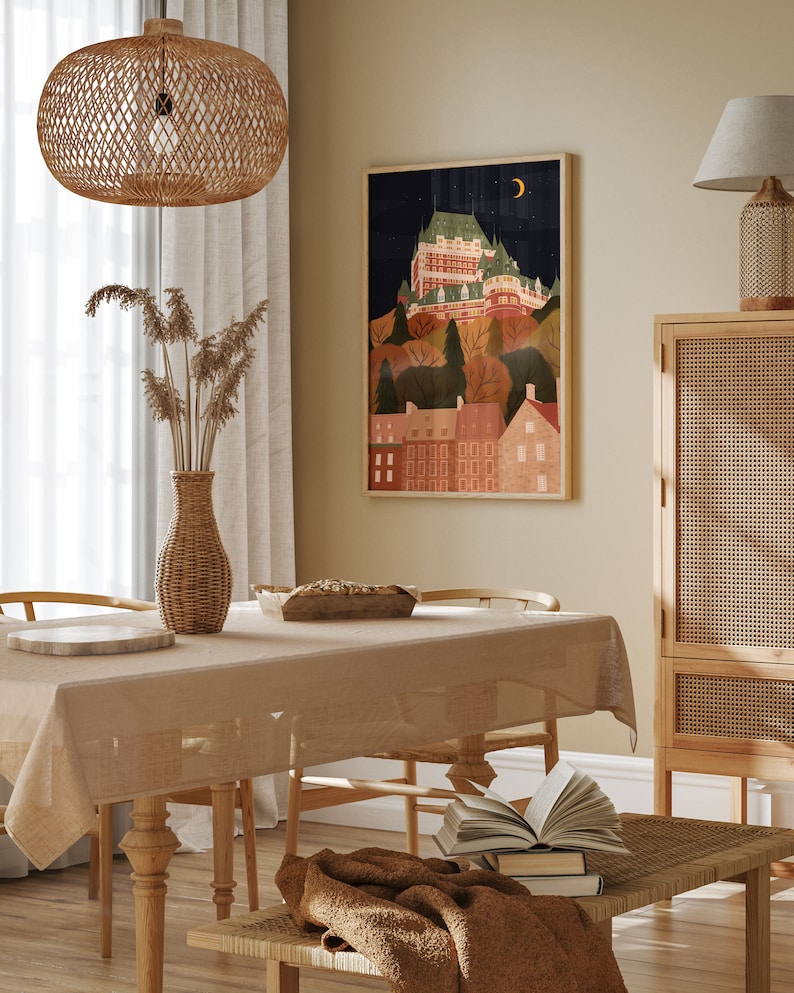 May include: A beige tablecloth covers a wooden dining table with two chairs. A brown throw blanket is draped over a wooden bench with a stack of books. A framed illustration of a city skyline with a crescent moon hangs on the wall above the table. A wicker lampshade hangs from the ceiling above the table. A wicker vase with dried flowers sits on the table.