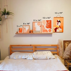 May include: A bedroom interior with a wooden bed frame and white bedding. The wall above the bed features a gallery wall of five abstract art prints in various sizes, including a print of a woman in a peach-colored dress. The prints are framed in natural wood and have a minimalist aesthetic. The wall also features a hanging plant in a macrame planter.