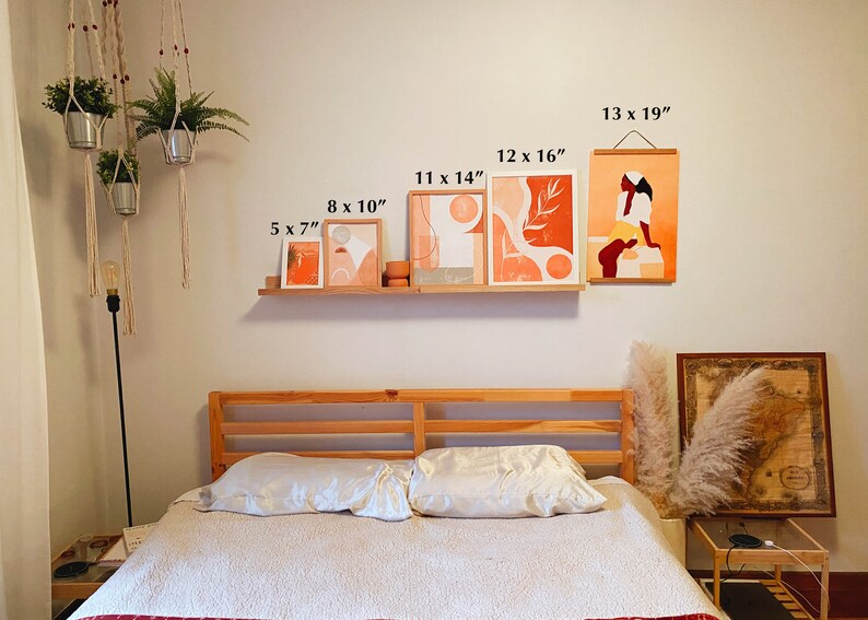 May include: A bedroom interior with a wooden bed frame and white bedding. The wall above the bed features a gallery wall of five abstract art prints in various sizes, including a print of a woman in a peach-colored dress. The prints are framed in natural wood and have a minimalist aesthetic. The wall also features a hanging plant in a macrame planter.