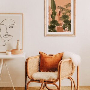 Puede incluir: Una silla de rat&aacute;n con un coj&iacute;n de piel sint&eacute;tica blanca y un coj&iacute;n de tiro naranja. La silla est&aacute; frente a una pared blanca con una impresi&oacute;n enmarcada de una mujer de pie en un jard&iacute;n tropical.