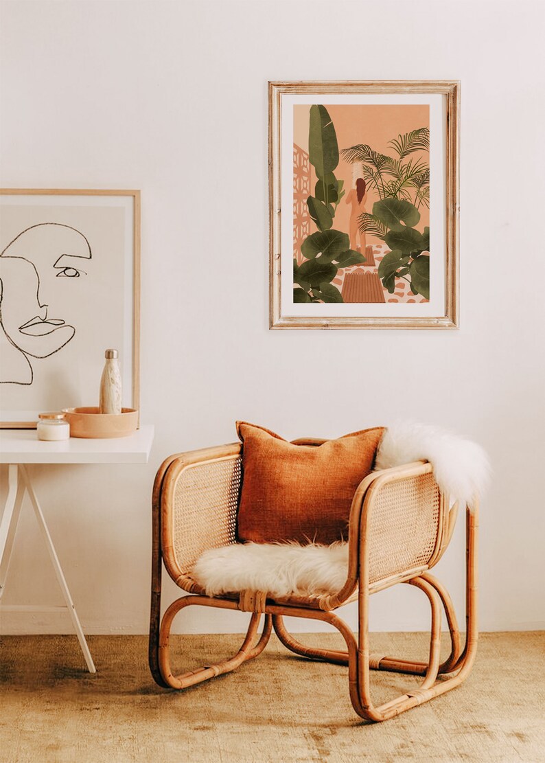 Puede incluir: Una silla de rat&aacute;n con un coj&iacute;n de piel sint&eacute;tica blanca y un coj&iacute;n de tiro naranja. La silla est&aacute; frente a una pared blanca con una impresi&oacute;n enmarcada de una mujer de pie en un jard&iacute;n tropical.