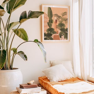 Puede incluir: Una ilustraci&oacute;n enmarcada de una mujer de pie en un jard&iacute;n tropical. La ilustraci&oacute;n est&aacute; en un marco de madera clara y est&aacute; colgada en una pared blanca. Una gran planta verde est&aacute; en una maceta blanca en primer plano. Un sof&aacute; cama blanco y naranja con almohadas est&aacute; en el fondo.