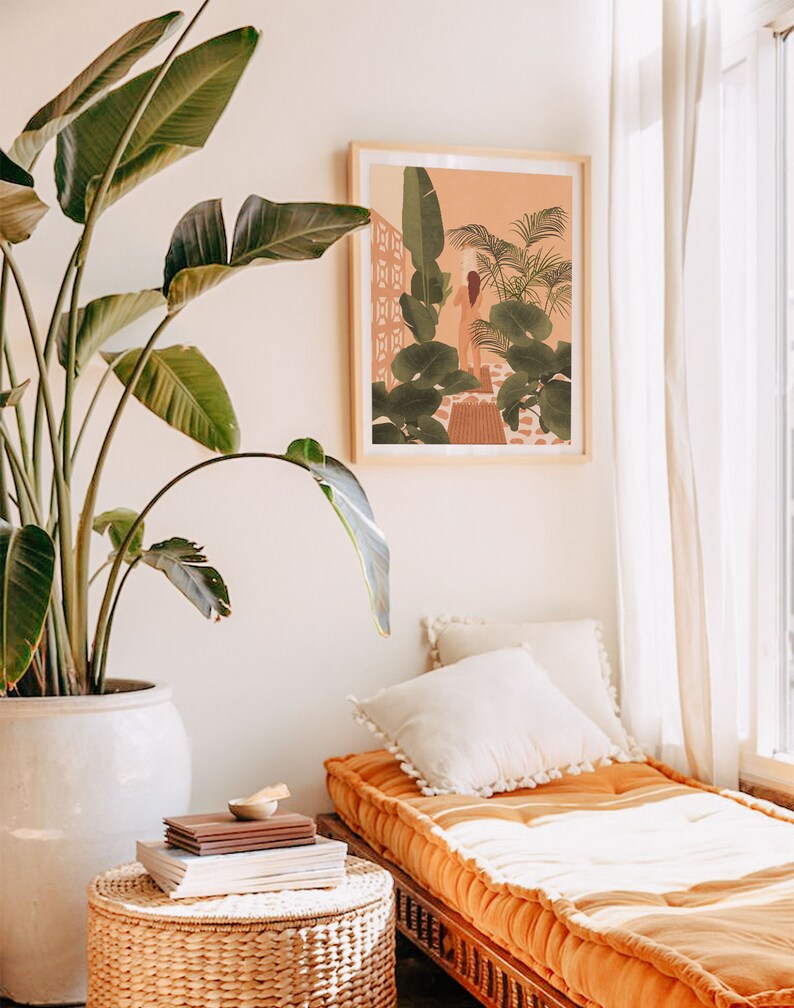 Puede incluir: Una ilustraci&oacute;n enmarcada de una mujer de pie en un jard&iacute;n tropical. La ilustraci&oacute;n est&aacute; en un marco de madera clara y est&aacute; colgada en una pared blanca. Una gran planta verde est&aacute; en una maceta blanca en primer plano. Un sof&aacute; cama blanco y naranja con almohadas est&aacute; en el fondo.