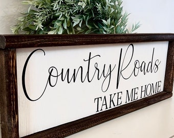 May include: A white wooden sign with a brown wood frame. The sign says "Country Roads Take Me Home" in black cursive lettering.
