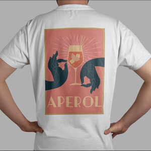 May include: A white t-shirt with a pink and orange graphic print of two hands holding a glass of Aperol Spritz. The text "APEROL" is printed in gold below the glass.