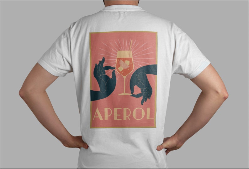 May include: A white t-shirt with a pink and orange graphic print of two hands holding a glass of Aperol Spritz. The text "APEROL" is printed in gold below the glass.