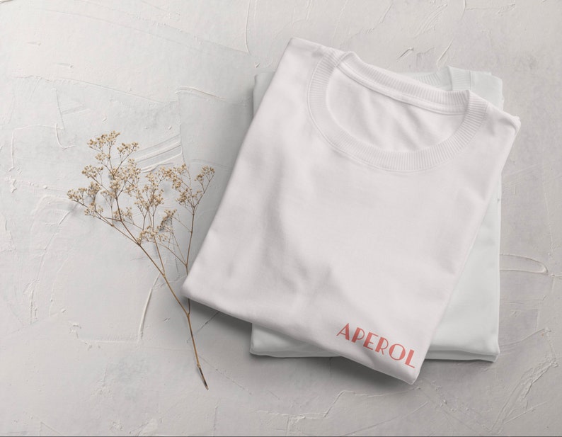 May include: White t-shirt with the word "APEROL" printed in pink on the front.