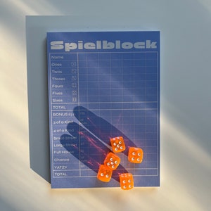 May include: A blue and white scorecard with the word "Spielblock" printed on it. There are six orange dice with white dots on a white surface.
