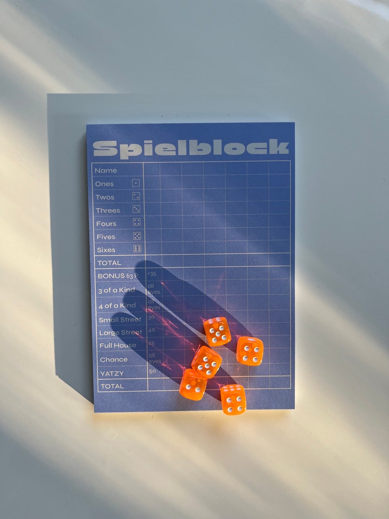 May include: A blue and white scorecard with the word "Spielblock" printed on it. There are six orange dice with white dots on a white surface.