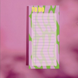 Könnte beinhalten: Ein vertikaler Notizblock vor einem rosa Hintergrund. Der Notizblock hat ein hellrosa Innere mit horizontalen Linien zum Schreiben. Oben auf dem Notizblock steht "TO DO" in Gelb. Der Notizblock hat ein grünes und weißes Wellenmuster an den Seiten.
