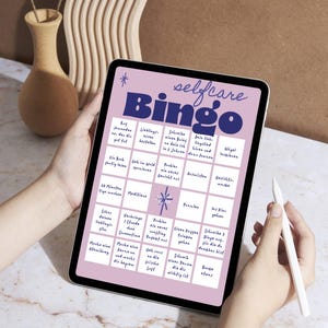 Può includere: Un tablet digitale che mostra un gioco "selfcare Bingo" con sfondo viola e testo bianco. Il tablet è tenuto da due mani, con una penna nella mano destra. Il gioco include varie attività di cura di sé.