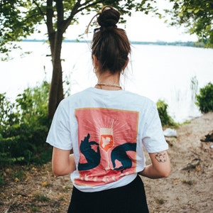 May include: A white t-shirt with a pink and orange graphic of a wine glass with two hands holding it. The text "Cheers" is visible on the graphic.