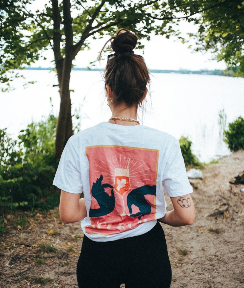 May include: A white t-shirt with a pink and orange graphic of a wine glass with two hands holding it. The text "Cheers" is visible on the graphic.