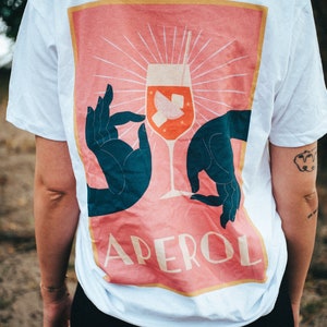 May include: White t-shirt with a pink and orange graphic of a cocktail glass with the word "APEROL" printed on it.