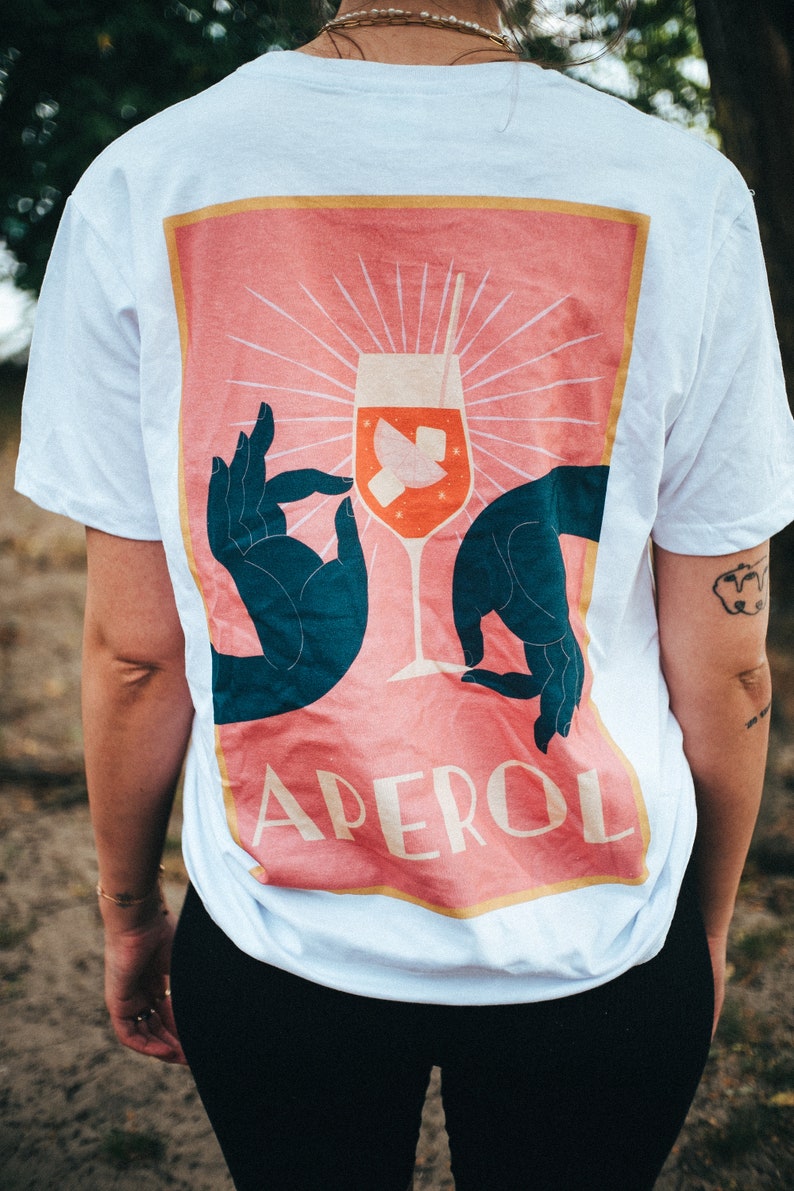 May include: White t-shirt with a pink and orange graphic of a cocktail glass with the word "APEROL" printed on it.