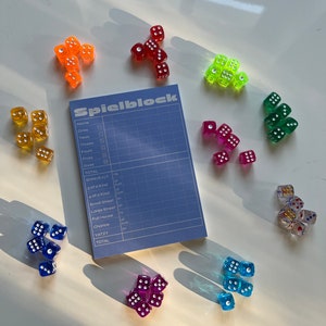 May include: A blue and white scorecard with the word "Spielblock" printed on it.  The scorecard is surrounded by colourful dice with white dots. The dice are red, orange, yellow, green, blue, purple, and gold.
