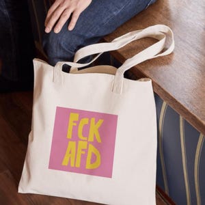 Puede incluir: Una bolsa de lona blanca con texto rosa y amarillo que dice "FCK AFD".