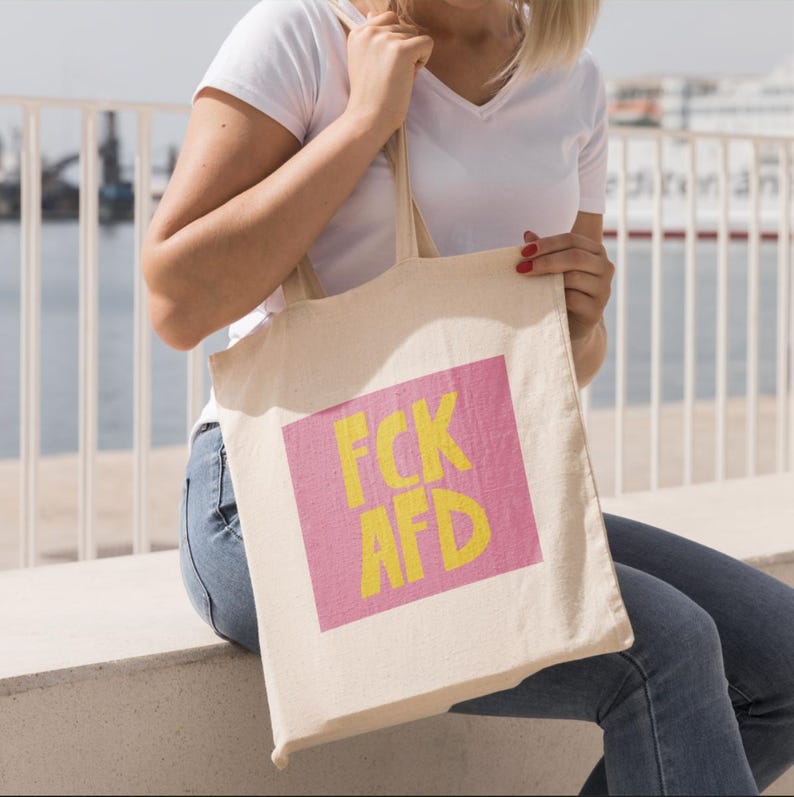 Puede incluir: Una persona sostiene una bolsa de tela blanca con un cuadrado rosa y el texto "FCK AFD" en letras amarillas.