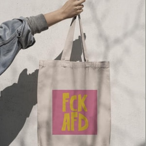 Puede incluir: Una bolsa de tela beige con un cuadrado rosa en la parte delantera. El cuadrado tiene el texto "FCK AFD" en letras amarillas.