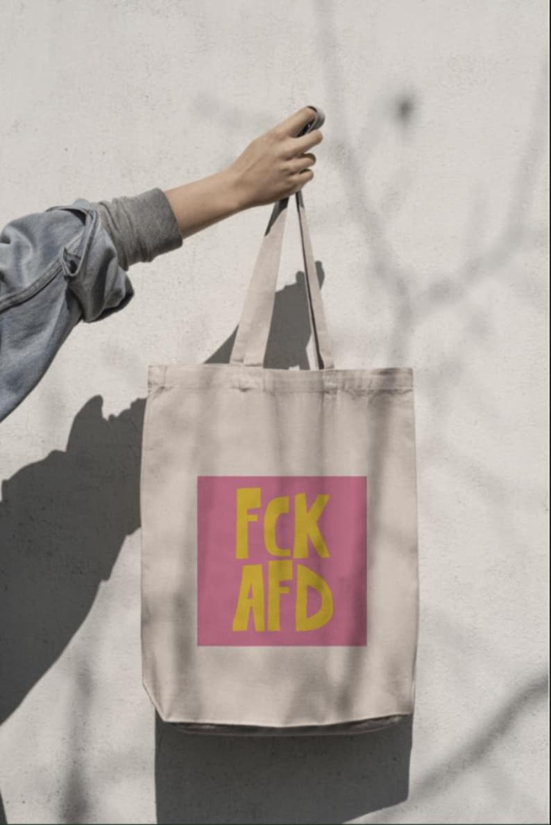 Puede incluir: Una bolsa de tela beige con un cuadrado rosa en la parte delantera. El cuadrado tiene el texto "FCK AFD" en letras amarillas.