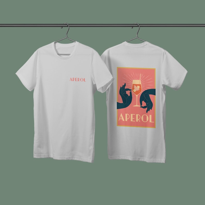 May include: Two white t-shirts with short sleeves. The front t-shirt has the word "APEROL" printed in black. The back t-shirt has a graphic of a hand holding a glass of Aperol Spritz with the word "APEROL" printed in black below the graphic.
