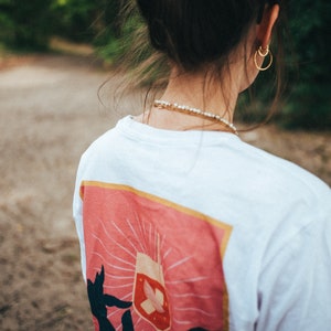 May include: White t-shirt with a pink graphic of a hand holding a wine glass with a butterfly inside. The graphic has a yellow border.