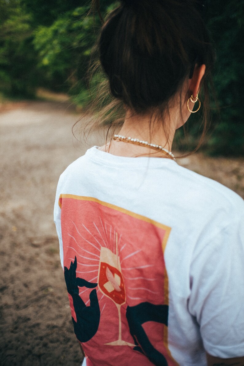 May include: White t-shirt with a pink graphic of a hand holding a wine glass with a butterfly inside. The graphic has a yellow border.