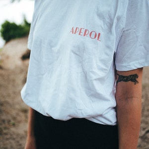 May include: A white t-shirt with the word "APEROL" printed in red on the front. The shirt is being worn by a person with a tiger tattoo on their arm.