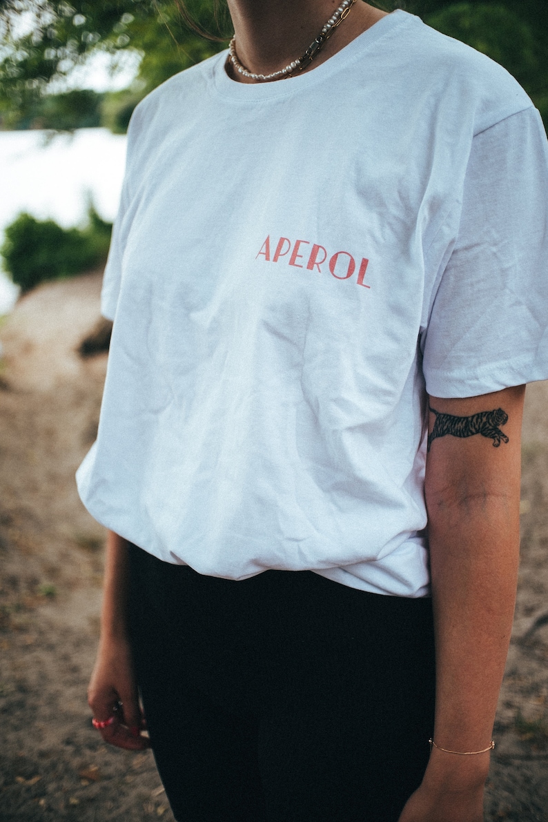 May include: A white t-shirt with the word "APEROL" printed in red on the front. The shirt is being worn by a person with a tiger tattoo on their arm.