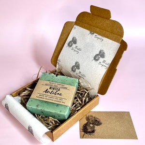 A small brown gift box containing a green bar of soap. The soap is labelled "Mintea Antibac" and is made with green tea leaves and peppermint essential oil. The box is lined with white thistle paper and has a small brown card with a thistle design on it.