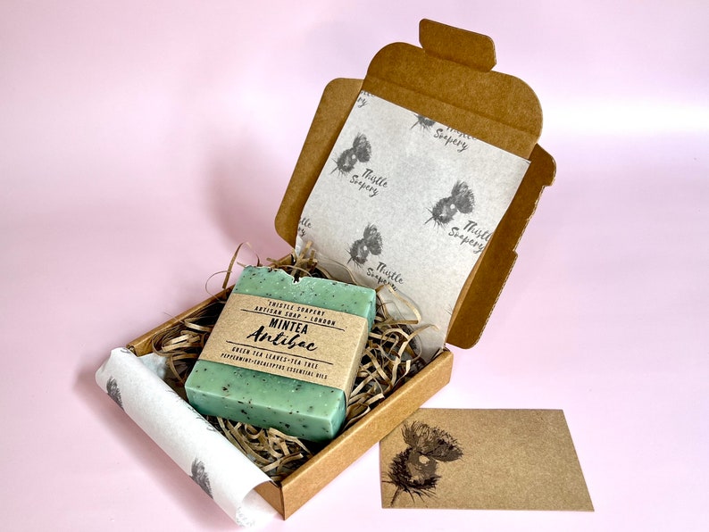 A small brown gift box containing a green bar of soap. The soap is labelled "Mintea Antibac" and is made with green tea leaves and peppermint essential oil. The box is lined with white thistle paper and has a small brown card with a thistle design on it.