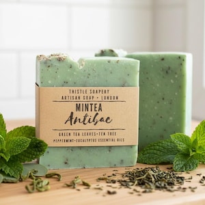 K&ouml;nnte beinhalten: Ein rechteckiges St&uuml;ck hellgr&uuml;ne Seife mit sichtbaren dunklen Sprenkeln, umwickelt mit einem beigen Papieretikett mit der Aufschrift Thistle Soapery Artisan Soap London Mintea Antibac Green Tea Leaves Tea Tree Peppermint-Eucalyptus Essential Oils.