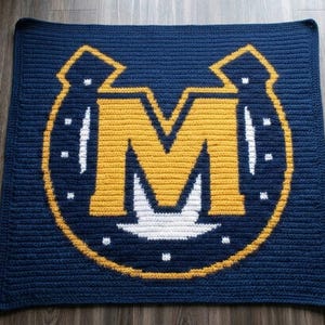May include: A square navy blue crochet blanket featuring a large yellow letter M centered within a yellow horseshoe outline. The design includes white accents within the horseshoe shape and small white dots arranged around the central letter M.