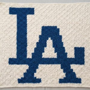 May include: A handmade, square crochet blanket featuring the letters "LA" in a bold, dark blue against a cream-colored background. The blanket is crafted with a textured stitch, showcasing the letters in a blocky, pixelated style.
