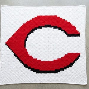 Könnte beinhalten: Eine weiße gehäkelte Decke mit einem großen roten Buchstaben "C" mit schwarzer Umrandung. Die quadratische Decke hat eine strukturierte Oberfläche. Der Buchstabe "C" ist das Hauptelement des Designs und nimmt den größten Teil der Fläche ein.